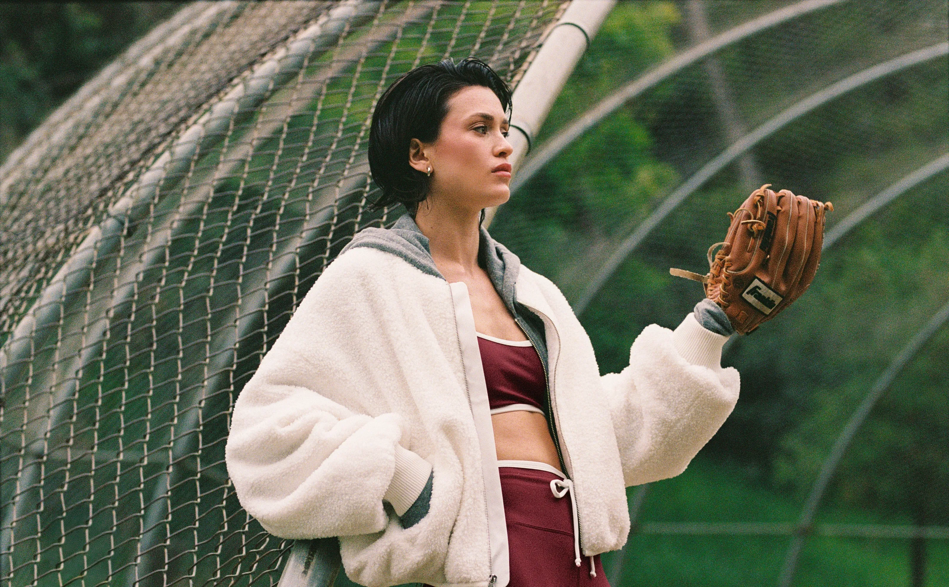 A model in a cream fleece bomber holding a baseball glove, against a backstop net